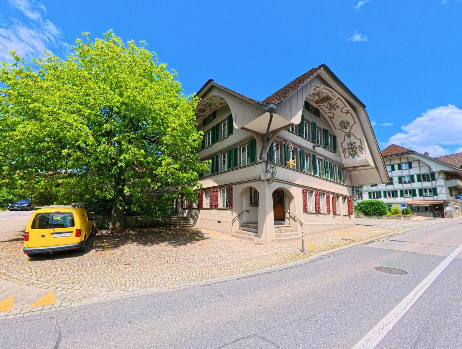 Vor einem historischen Gebäude mit bemalten Fassaden in Grosshoechstetten (3506, Kanton Bern) steht ein gelber Lieferwagen unter einem grossen, belaubten Baum.