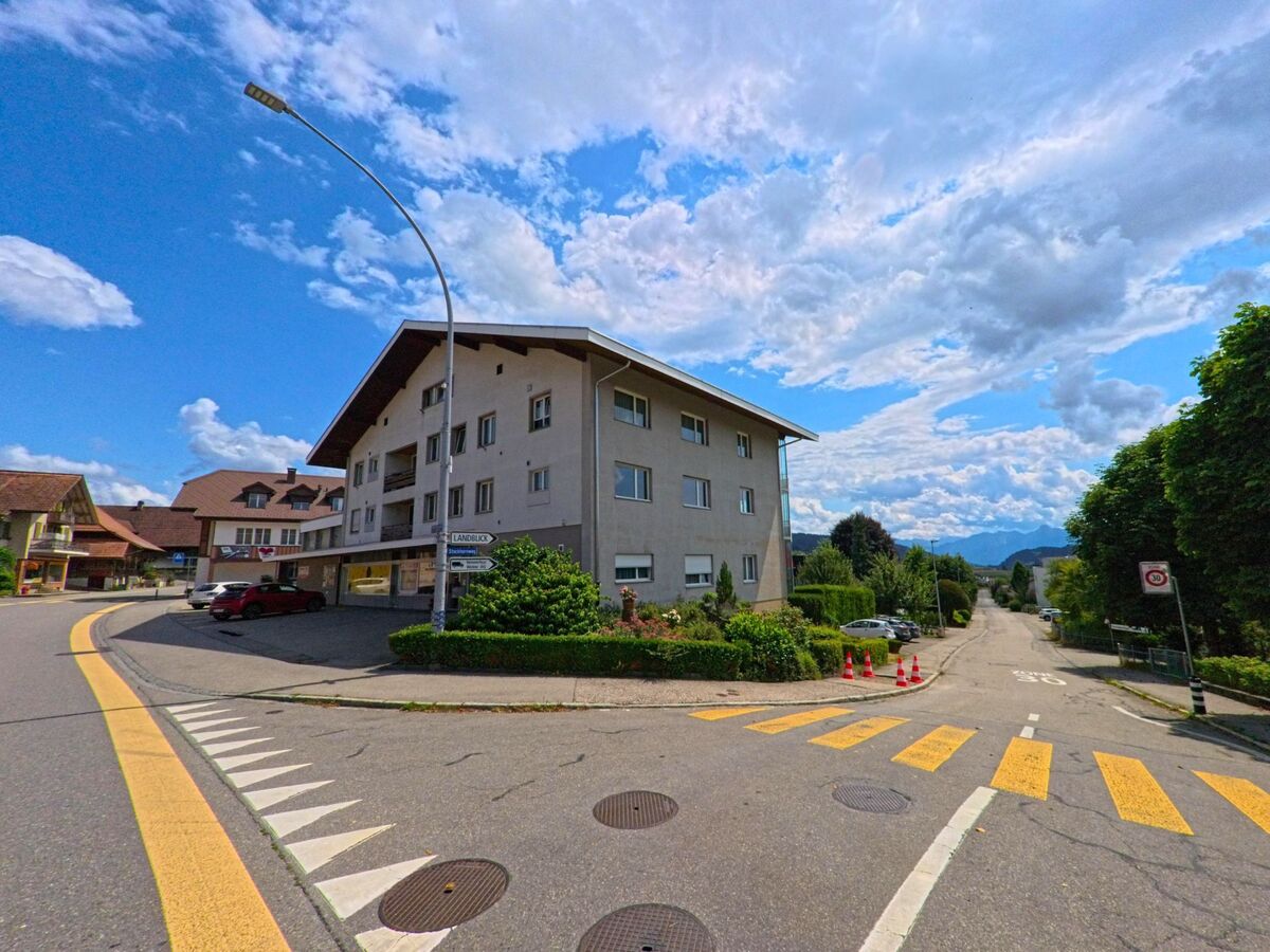 In Grosshoechstetten (3506, Kanton Bern) zweigt eine verkehrsberuhigte Seitenstrasse mit Blick auf die Hügel vom belebten Kreuzungsbereich mit mehrgeschossigen Wohnbauten ab.