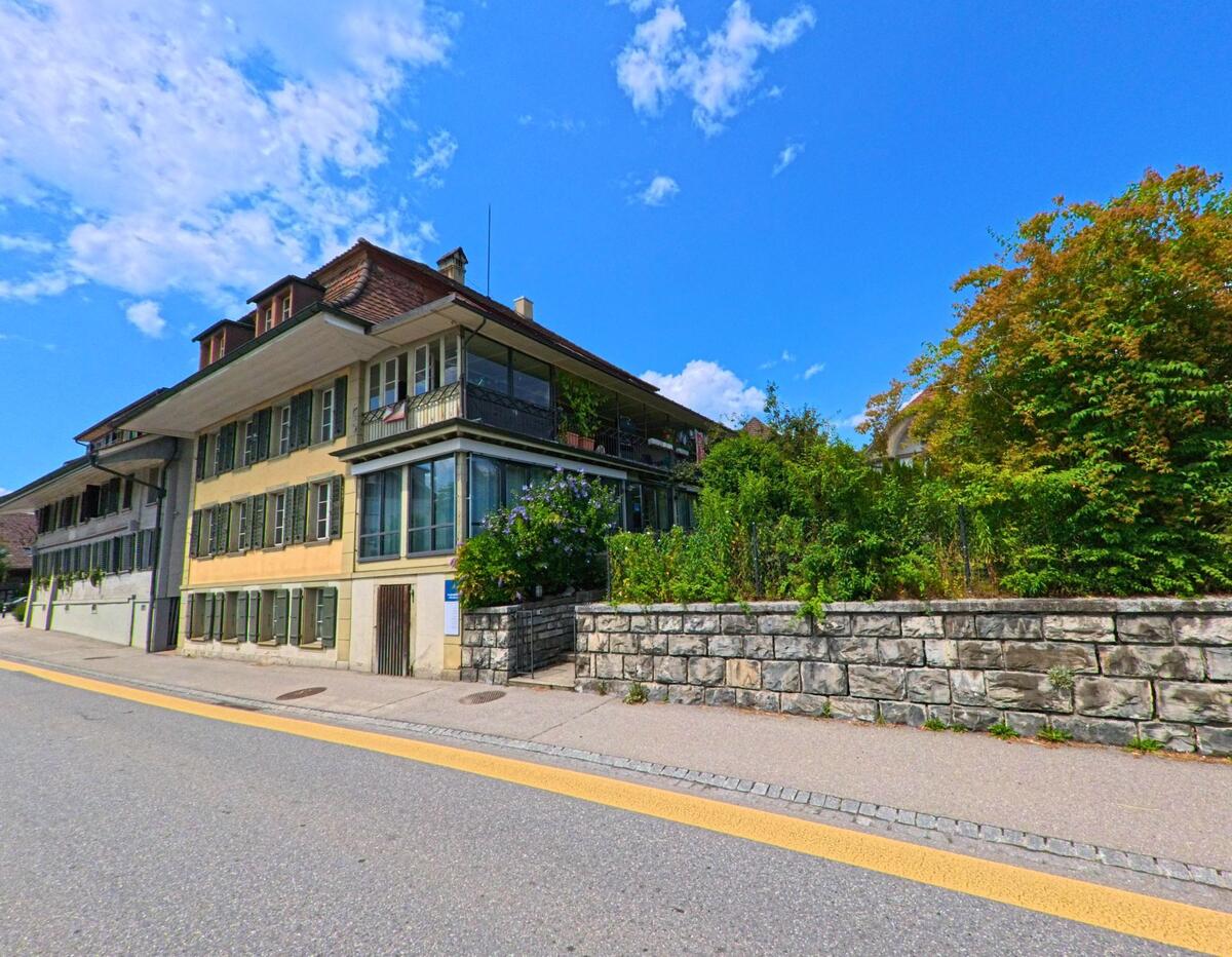 In Grosshoechstetten (3506, Kanton Bern) steht ein grosses Wohnhaus mit gruenen Fensterlaeden und verglastem Wintergarten direkt an einer leicht ansteigenden Strasse neben dichter Gartenbepflanzung.