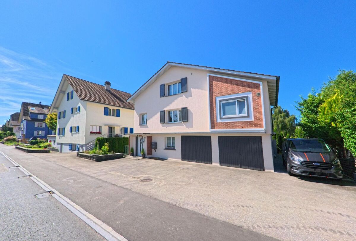 In Grosswangen (6022, Kanton Luzern) reihen sich entlang einer Quartierstrasse mehrere Einfamilienhaeuser mit Garagen und kleinen Vorgartenbereichen.