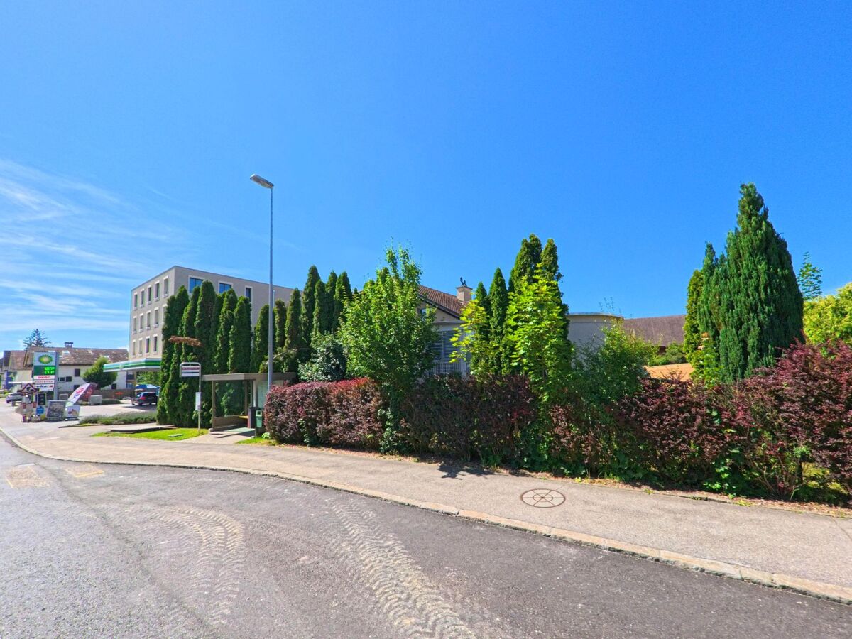 In Grosswangen (6022, Kanton Luzern) liegt eine moderne Tankstelle mit angrenzendem Geschaeftsgebaeude neben einer Bushaltestelle und dicht bepflanztem Strassenrand mit Hecken und Zypressen.