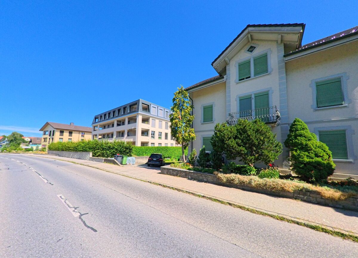 In Grosswangen (6022, Kanton Luzern) stehen ein Wohnhaus mit gruener Fensterlaeden und Zierbalkon sowie moderne Mehrfamilienhaeuser entlang einer leicht abschuessigen Strasse.