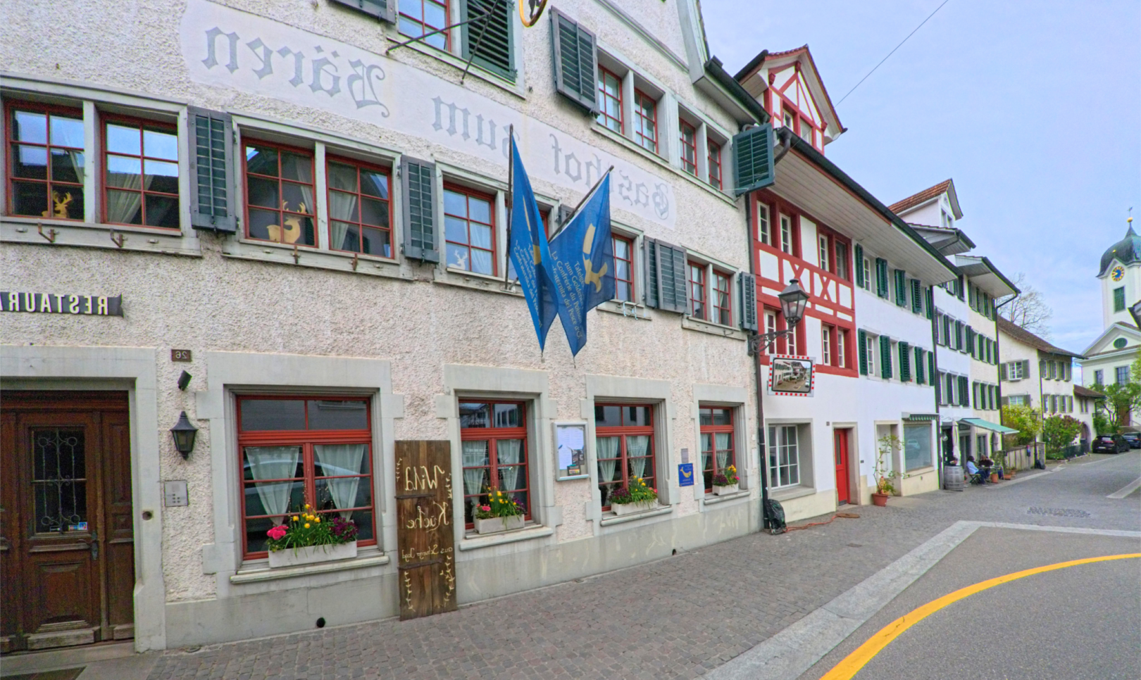 Im ORT Grüningen, PLZ 8627 im KANTON Zürich reiht sich ein traditionelles Gasthaus mit Fahnen und Sprossenfenstern in die historische Altstadtgasse nahe der Kirche ein.
