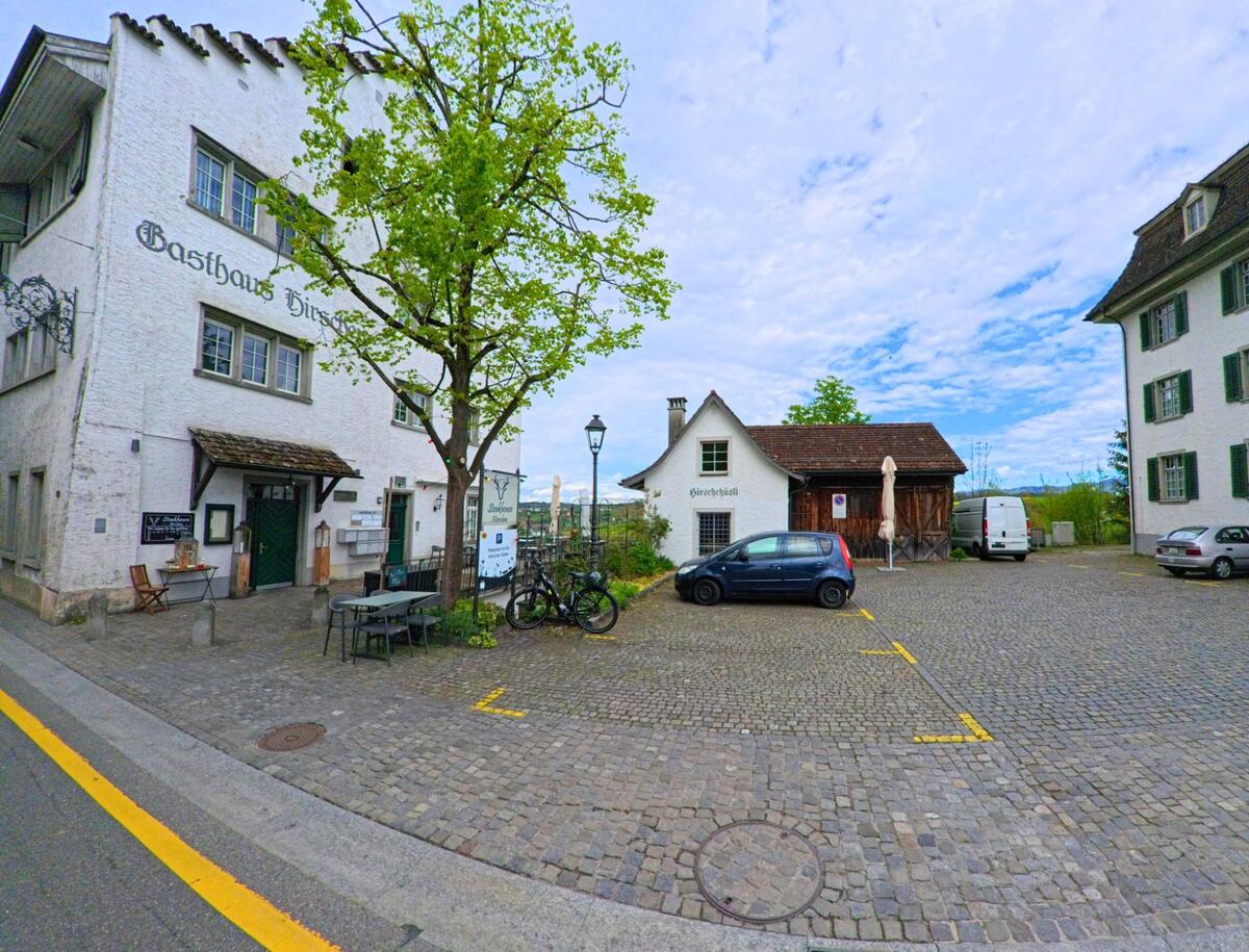 In Grüningen, PLZ 8627 im KANTON Zürich befindet sich das Gasthaus Hirschen an einem gepflasterten Platz mit Baum, Sitzgelegenheiten und angrenzendem Parkplatz.