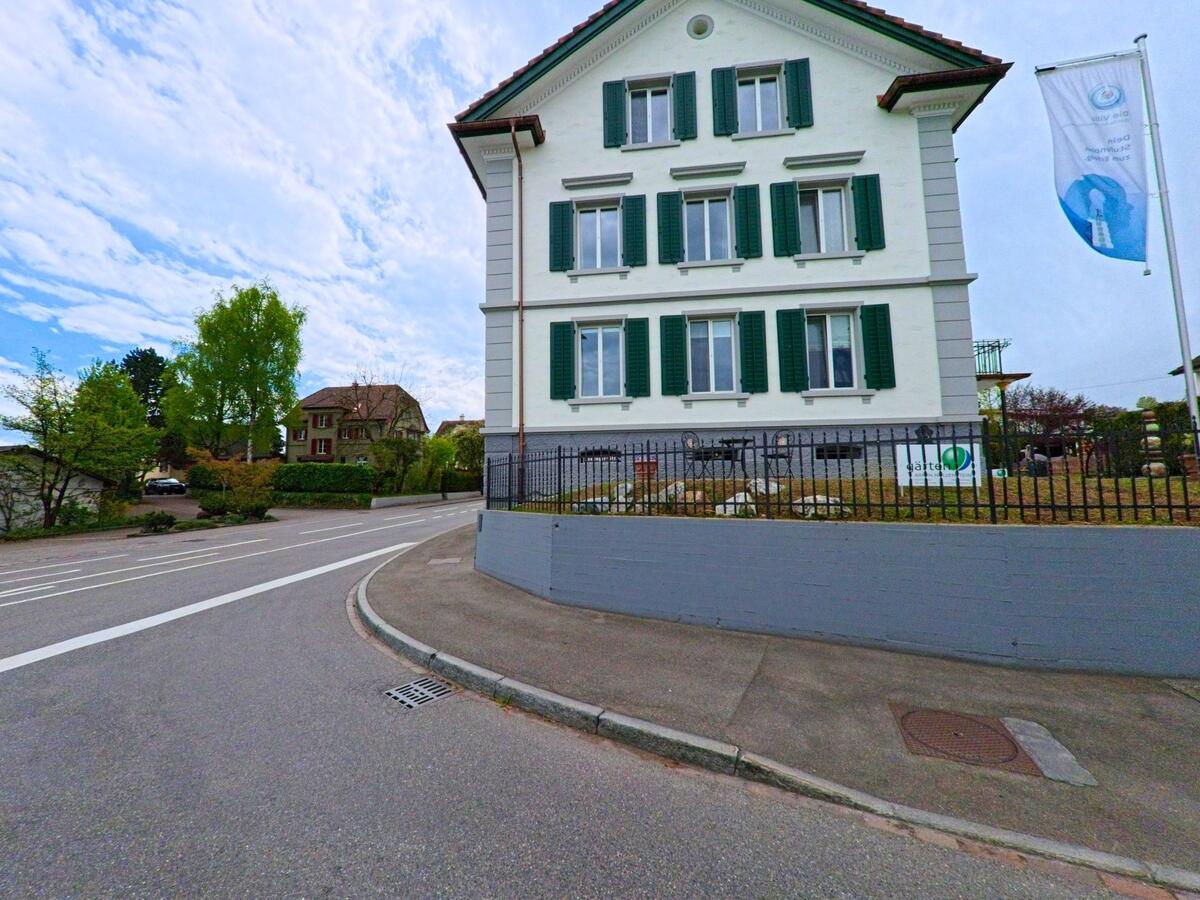 In Grüningen, PLZ 8627 im KANTON Zürich steht ein renoviertes Gebäude mit grünen Fensterläden und gepflegtem Vorplatz an einer sanft geschwungenen Strasse.