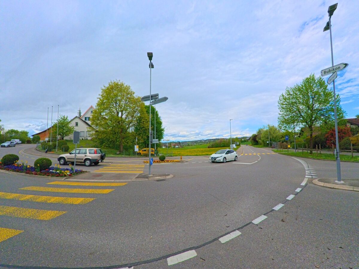 Am Ortsrand von Grüningen, PLZ 8627 im KANTON Zürich trifft eine mehrspurige Kreuzung mit Zebrastreifen und Wegweisern auf eine offene Landschaft mit Blick Richtung Bubikon.