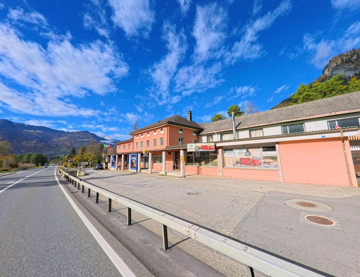 Entlang der Hauptstrasse in Grüsch (7213, Kanton Graubünden) stehen Geschäfts- und Wohnbauten mit Schaufenstern vor der offenen Berglandschaft.