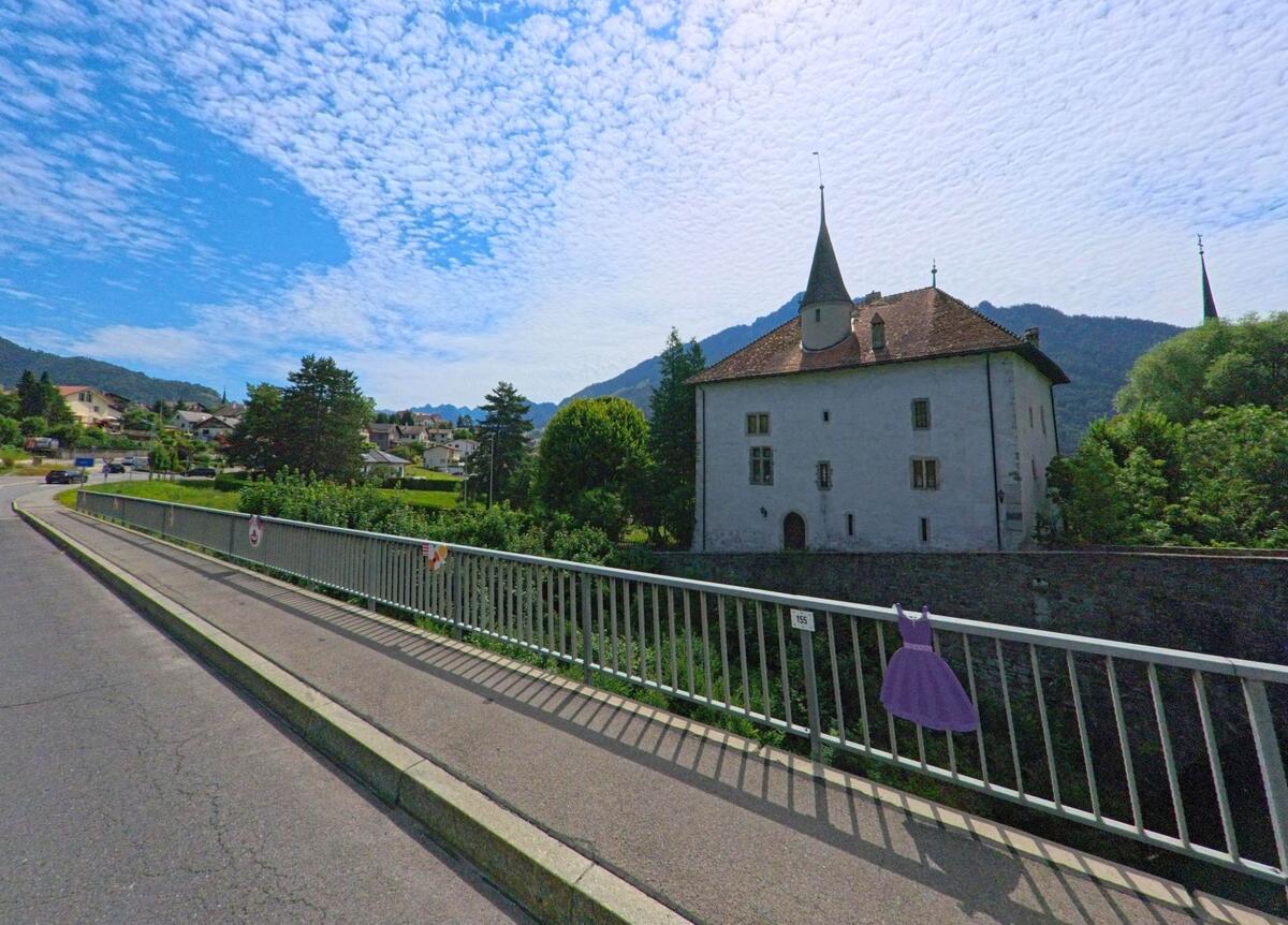 In Gruyères (1663, Kanton Freiburg) führt eine Brücke mit Geländer an einem historischen Gebäude mit Turm vorbei, umgeben von dichtem Grün und vor einer bewaldeten Hügellandschaft.