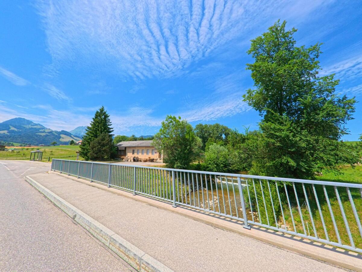 Eine kleine Brücke mit Metallgeländer in Gruyères (1663, Kanton Freiburg) überquert einen Bachlauf, im Hintergrund stehen ein helles Gebäude, Bäume und sanfte Hügel.