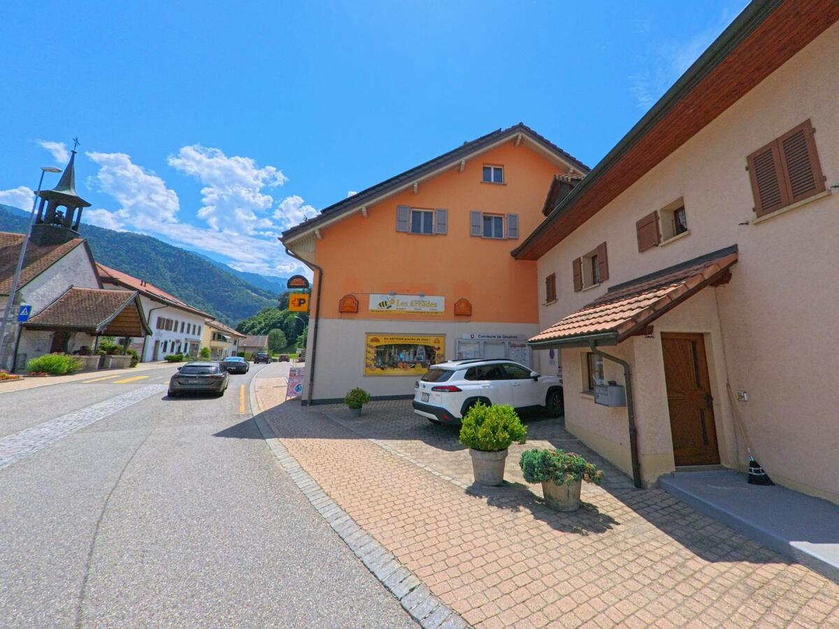 In Gruyères (1663, Kanton Freiburg) verläuft eine Dorfstrasse an einer Kirche mit Glockenturm vorbei, flankiert von traditionellen Häusern und einem orangefarbenen Geschäftsgebäude.
