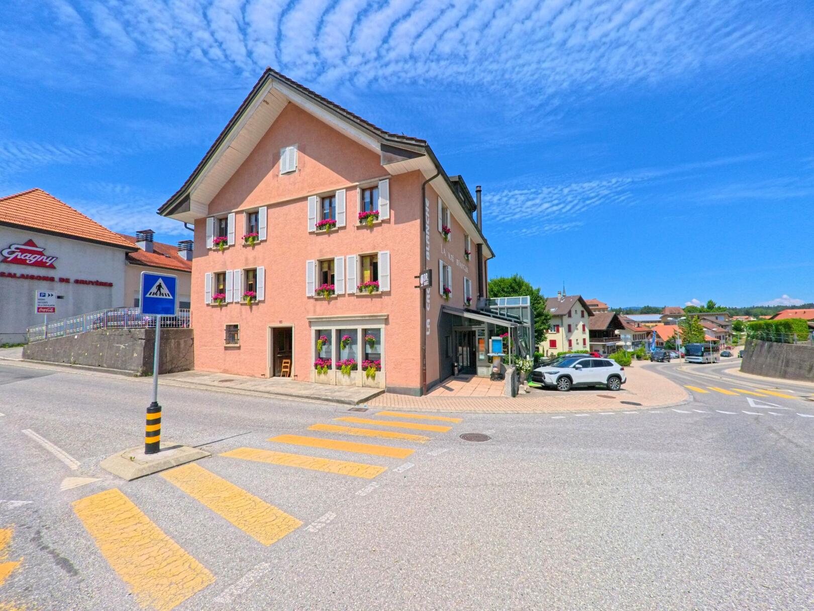 In Gruyères (1663, Kanton Freiburg) steht ein rosafarbenes Haus mit Fensterblumen an einer Kreuzung mit Zebrastreifen, umgeben von weiteren Gebäuden im Dorfzentrum.