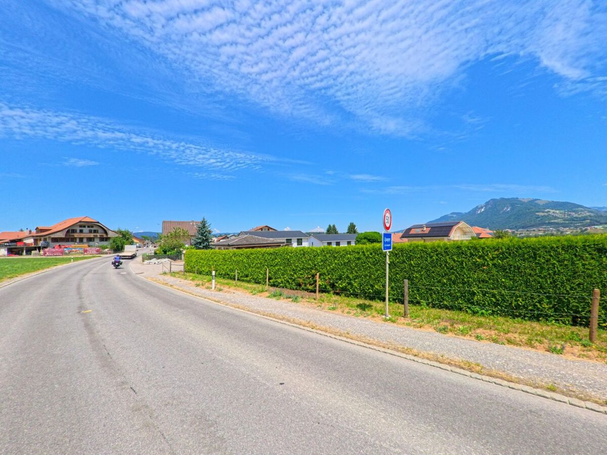 Am Ortseingang von Gruyères (1663, Kanton Freiburg) führt eine geschwungene Landstrasse an Wohnhäusern und einer gepflegten Hecke entlang, mit Blick auf das hügelige Umland.