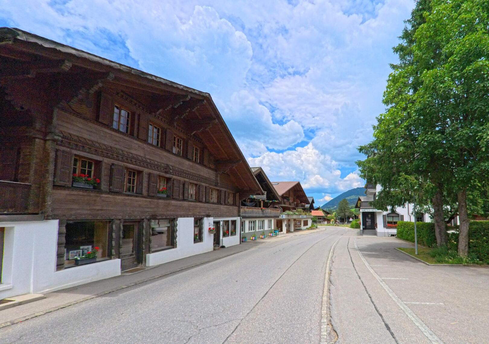 Entlang der leicht geschwungenen Hauptstrasse in Gsteig (3785, Kanton Bern) stehen traditionelle Holzhäuser mit Fensterläden und Blumenschmuck gegenüber einem schattigen Parkplatz.