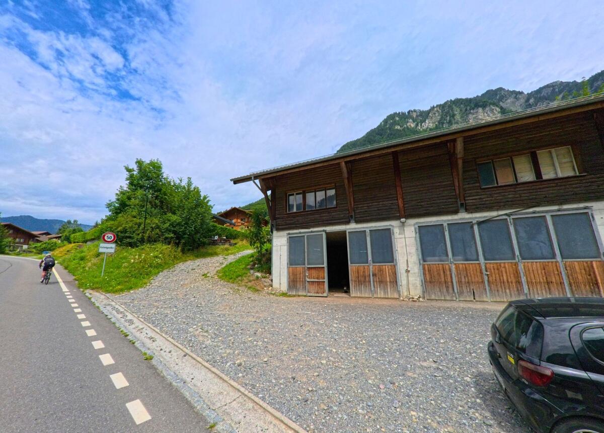 Am Ortseingang von Gsteigwiler (3814, Kanton Bern) passiert ein Velofahrer eine Geschwindigkeitsbegrenzung, während rechts ein grosser Holzbau mit Kiesfläche an der Strasse liegt.