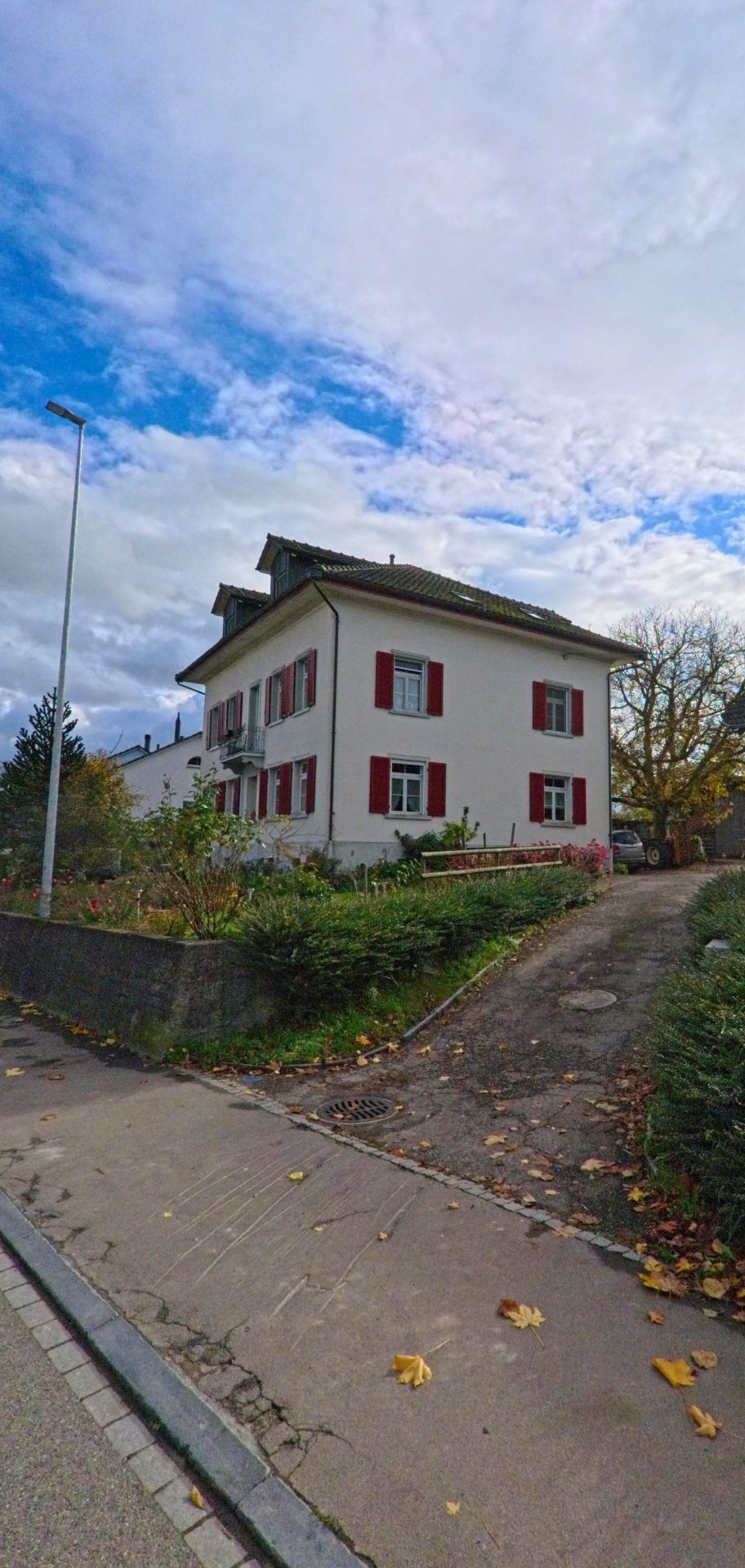 Ein Wohnhaus mit roten Fensterläden steht in Güttingen (8594, Kanton Thurgau) neben einem schmalen Zufahrtsweg und einem verwilderten Garten.