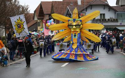 Eine farbenfrohe Fasnachtsgruppe mit grosser Figur zieht durch eine Strasse waehrend eines Umzugs in Riggisberg (3132, Kanton Bern).
