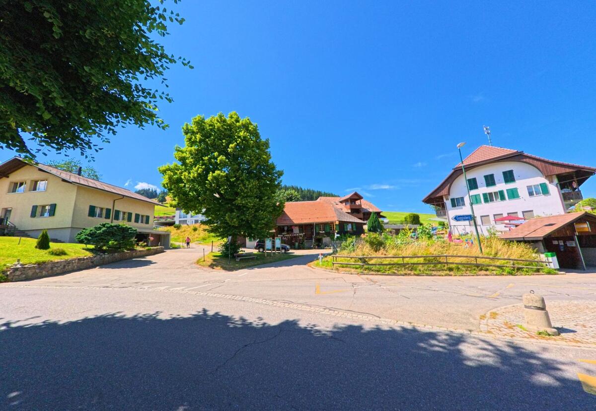 Im Zentrum von Guggisberg (3156, Kanton Bern) stehen mehrere Wohnhäuser mit traditionellen Fassaden vor einer grünen Hügellandschaft.