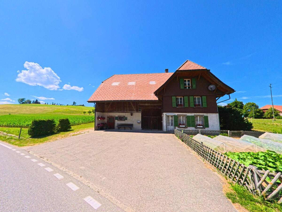 Ein Bauernhaus mit grün gestrichenen Fensterläden steht in Guggisberg (3158, Kanton Bern) am Rand eines gepflasterten Vorplatzes vor weiten Feldern und leicht hügeliger Landschaft.