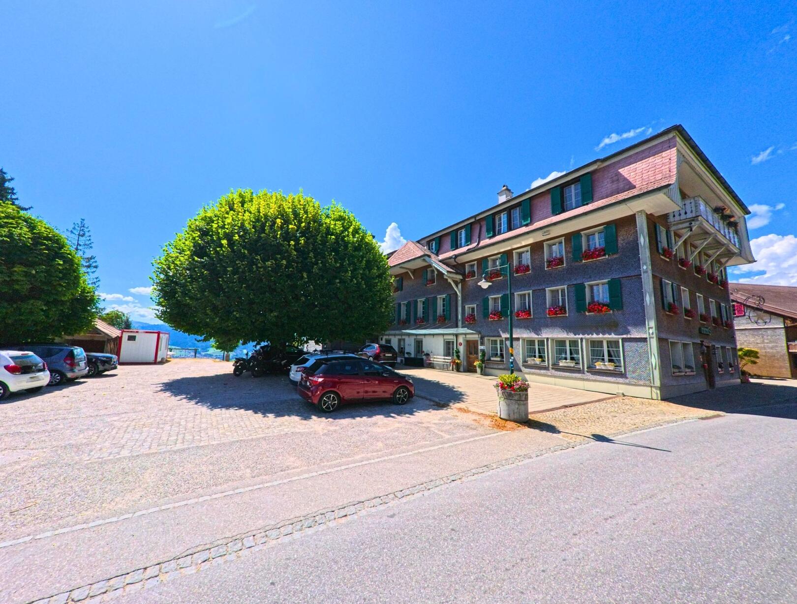 Vor einem Gasthof mit grün gestrichenen Fensterläden in Guggisberg (3158, Kanton Bern) erstreckt sich ein grosser Parkplatz unter schattenspendenden Bäumen.