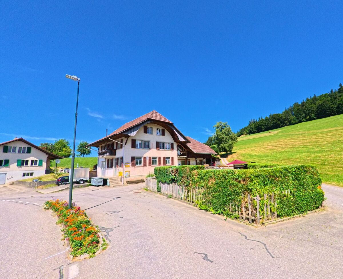 Am Rand von Guggisberg (3158, Kanton Bern) stehen mehrere Wohnhäuser vor offenen Wiesenflächen, begleitet von einer bunt bepflanzten Verkehrsinsel.
