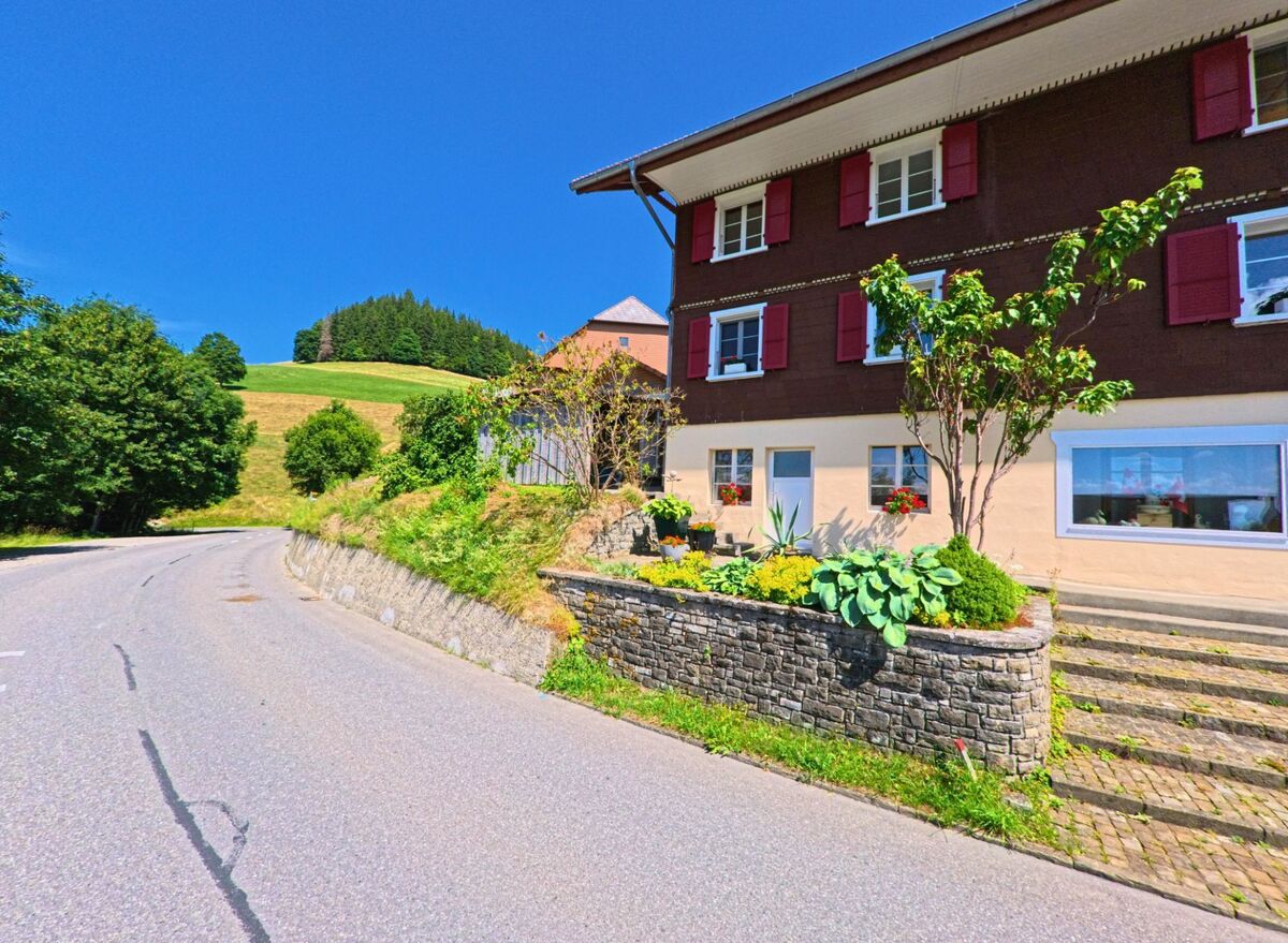 Entlang einer kurvigen Strasse in Guggisberg (3158, Kanton Bern) liegt ein Wohnhaus mit rotem Fensterladen und gepflegtem Vorgarten, umgeben von hügeliger Landschaft.