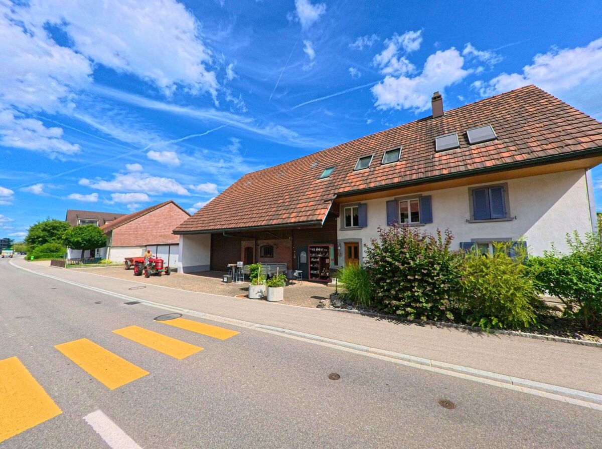 Ein Bauernhaus mit Verkaufsstand und rotem Traktor steht in Gunzgen (4617, Kanton Solothurn) direkt an einer Strasse mit Zebrastreifen unter einem strahlend blauen Himmel.