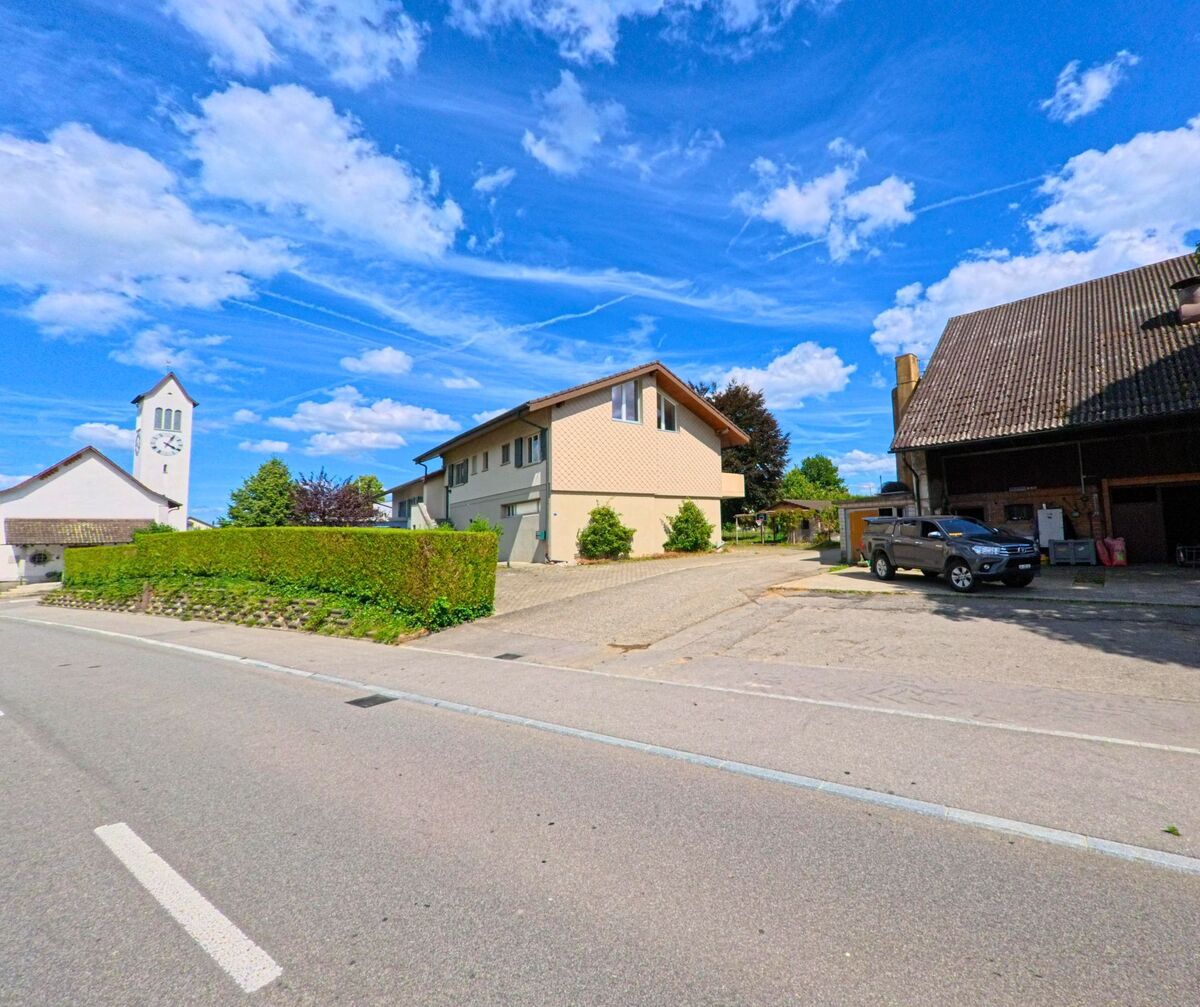 In Gunzgen (4617, Kanton Solothurn) erhebt sich der weisse Kirchturm mit Uhr hinter einem Wohnhaus und einem Bauernhof an einer ruhig gelegenen Strasse.