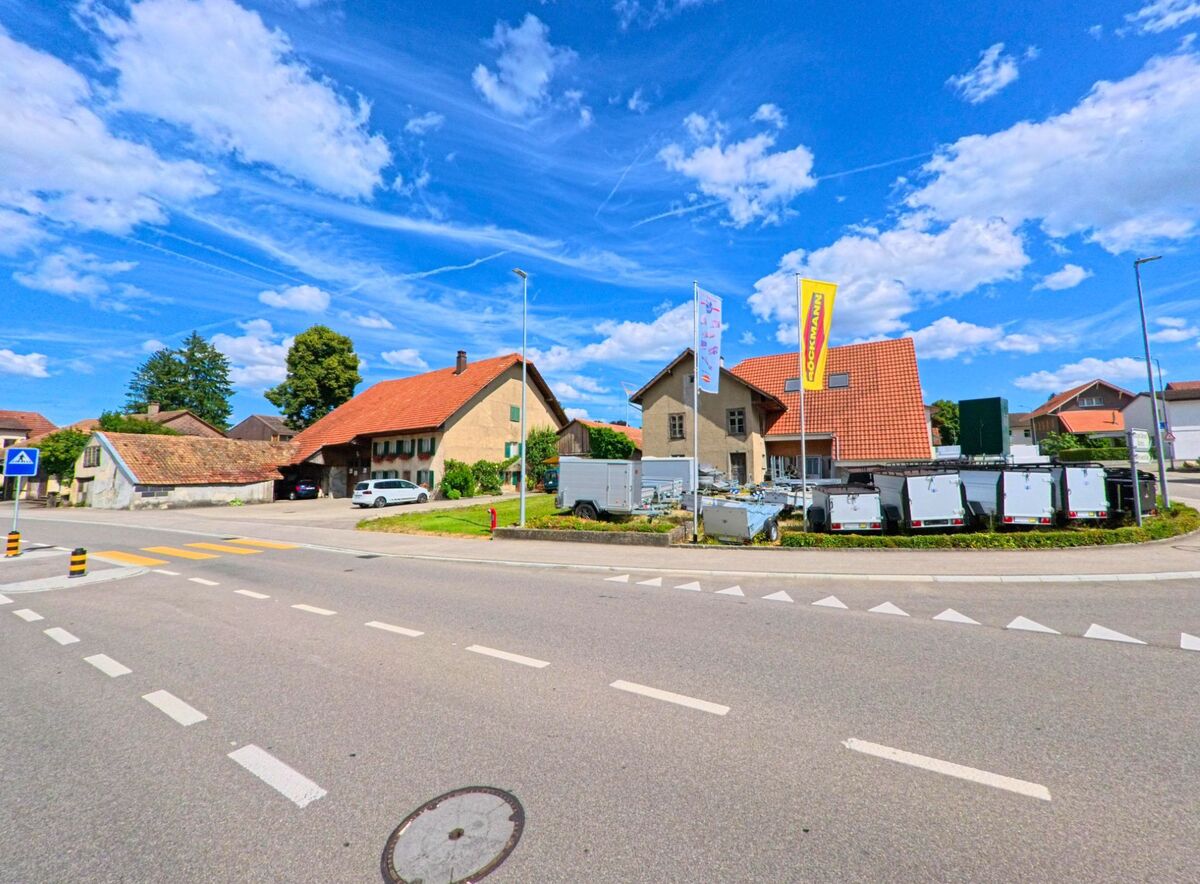 An einer Kreuzung in Gunzgen (4617, Kanton Solothurn) befinden sich ein Verkaufsgelaende mit Anhängern und mehrere Wohnhaeuser mit roten Daechern unter einem wolkigen Sommerhimmel.