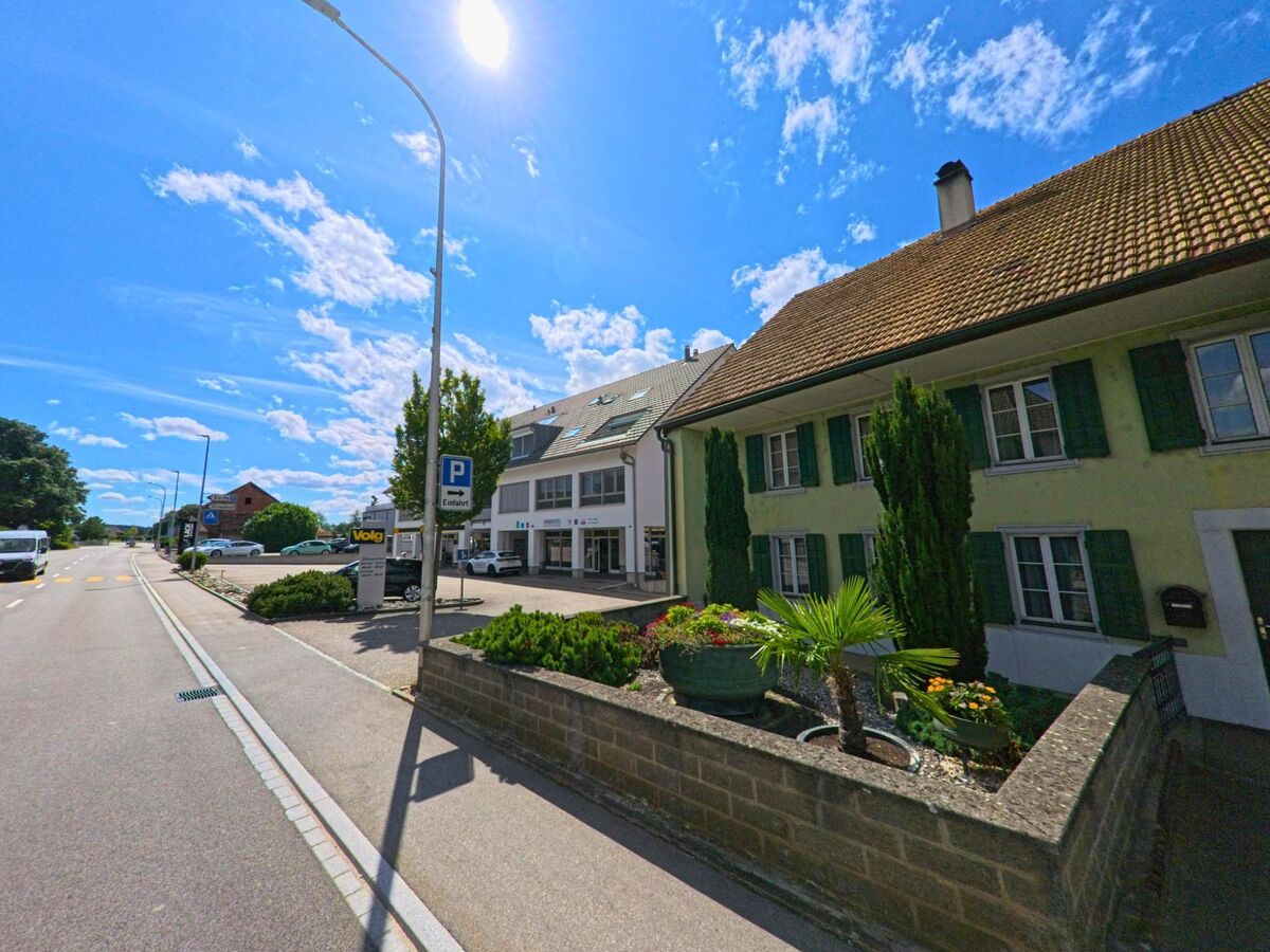 An der Hauptstrasse in Gunzgen (4617, Kanton Solothurn) stehen ein traditionelles Wohnhaus mit gruener Fassade und ein modernes Geschaeftsgebaeude mit Volg-Filiale und Parkplatz.