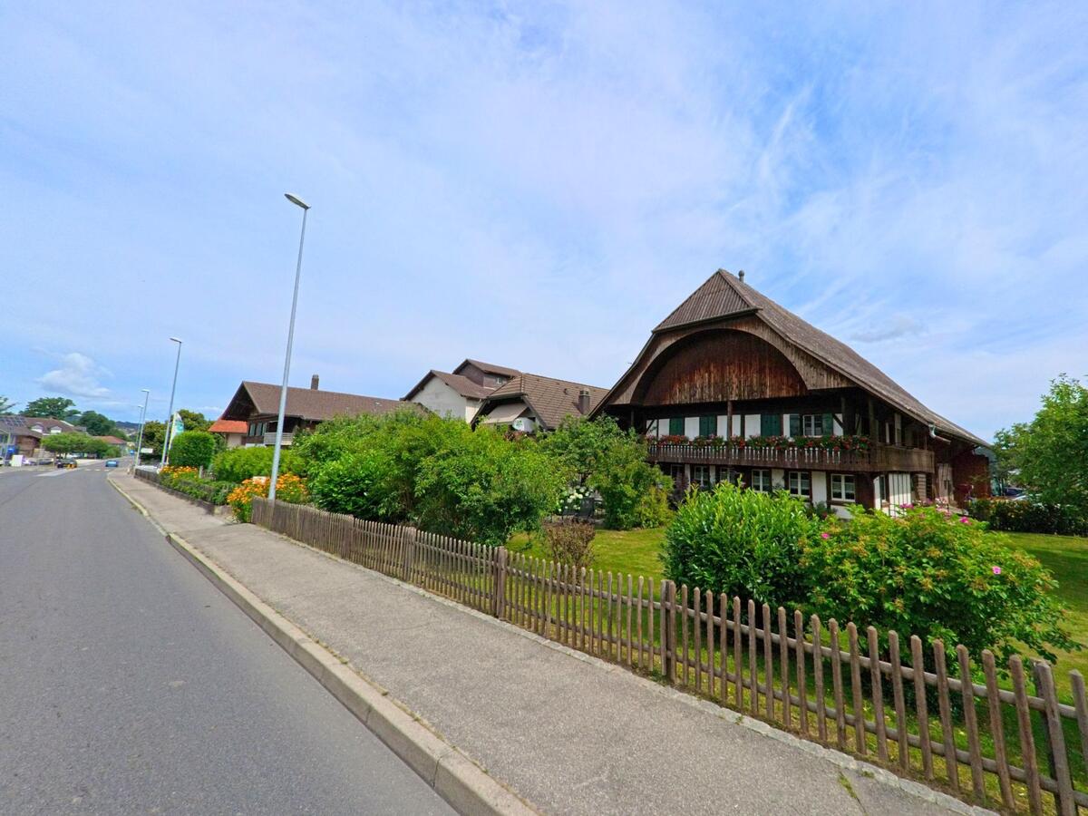 An einer Hauptstrasse in Gurmels (3212, Kanton Freiburg) steht ein traditionelles Bauernhaus mit weitgezogenem Dach und gepflegtem Garten hinter einem Holzzaun.