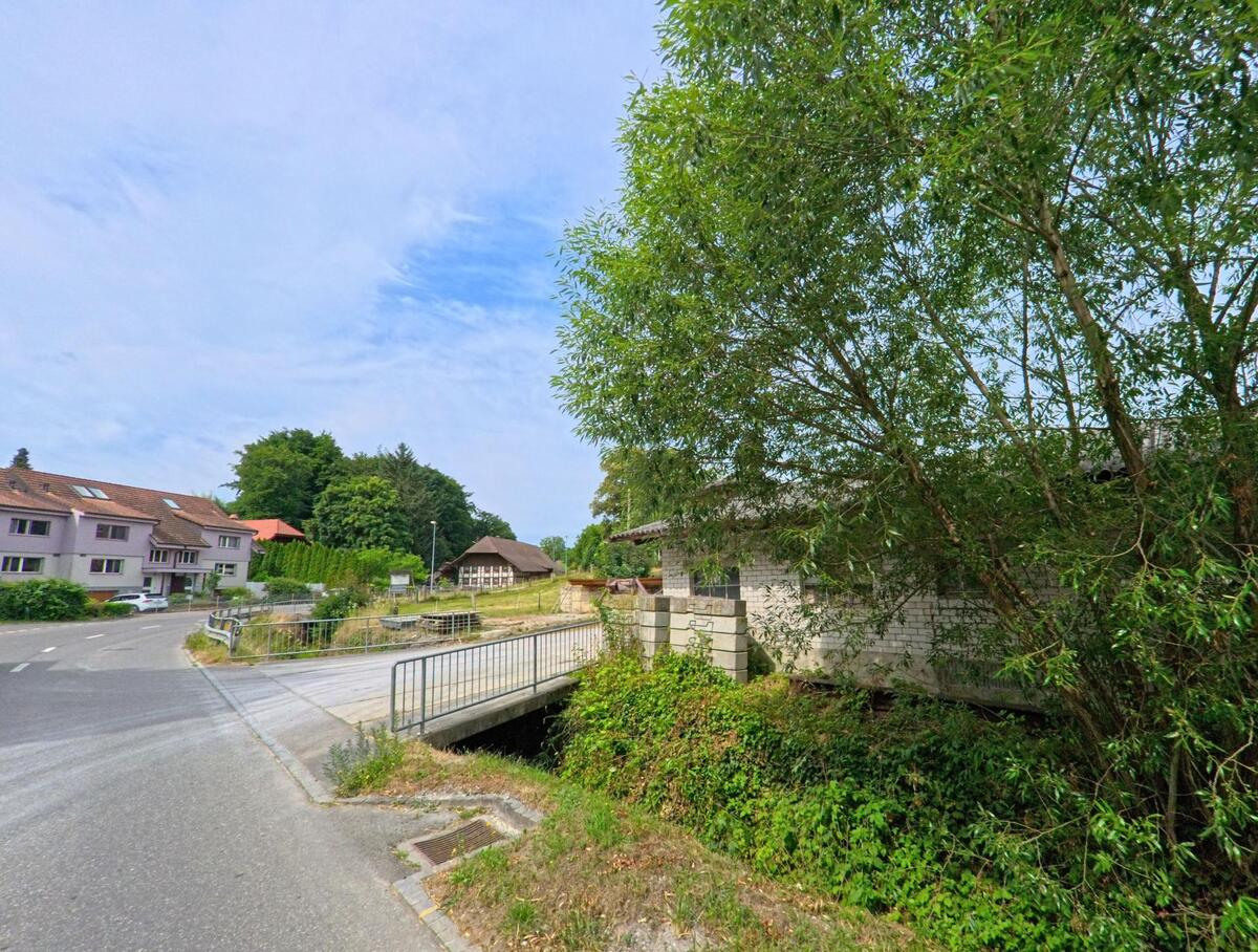 In Gurmels (3212, Kanton Freiburg) überspannt eine kleine Brücke einen Bachlauf und verbindet ein Wohngebiet mit einer leicht ansteigenden Strasse zu Bauernhäusern.