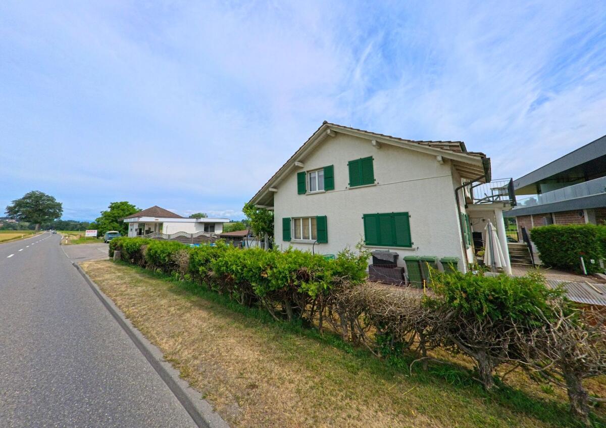 Am Ortsrand von Gurmels (3212, Kanton Freiburg) steht ein Einfamilienhaus mit grünen Fensterläden und Gartenbereich direkt an einer ruhig verlaufenden Landstrasse.