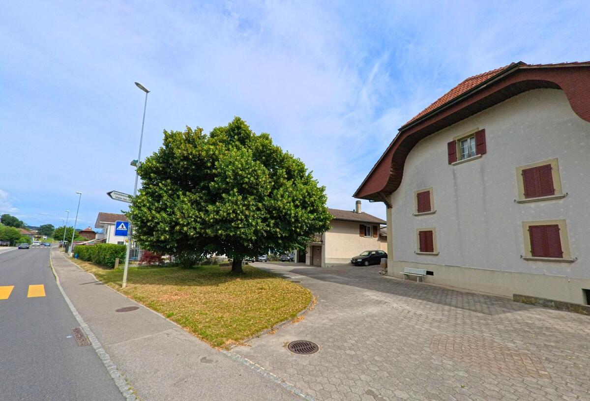 In Gurmels (3212, Kanton Freiburg) steht ein Haus mit geschwungenem Dach und roten Fensterläden an einer verkehrsberuhigten Strasse mit Fussgängerstreifen und Bauminsel.