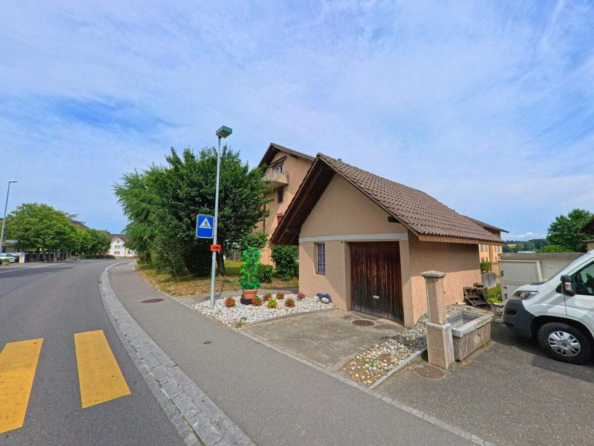 An einer Strasse in Gurmels (3212, Kanton Freiburg) steht ein kleines Haus mit Holztor neben einem Brunnen, während ein Fussgängerstreifen zur sicheren Querung führt.