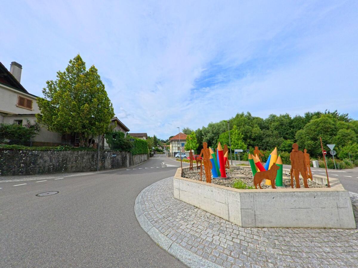 Ein Kreisverkehr in Gurmels (3212, Kanton Freiburg) ist mit farbigen Bleistift-Skulpturen und Figuren aus rostigem Metall gestaltet, umgeben von Wohnhäusern und Bäumen.