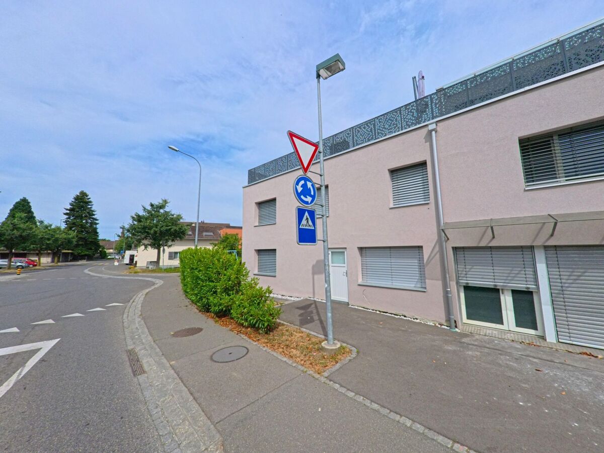  An einer Strassenecke in Gurmels (3212, Kanton Freiburg) steht ein modernes Gebäude mit geschlossenen Rollläden, daneben befinden sich Verkehrszeichen für einen nahen Kreisverkehr und Fussgängerstreifen.