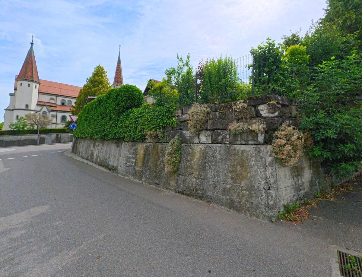 In Gurmels (3212, Kanton Freiburg) führen enge Strassen an einer hohen Stützmauer mit Begrünung vorbei, während im Hintergrund die markanten Kirchtürme emporragen.