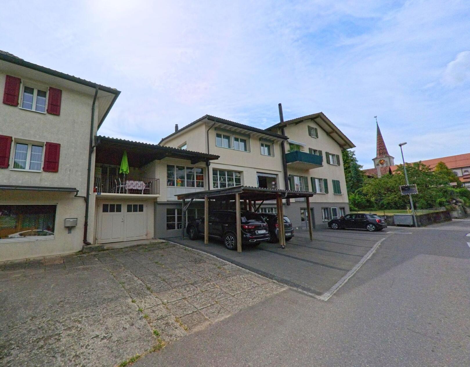 Im Zentrum von Gurmels (3212, Kanton Freiburg) stehen mehrere Wohnhäuser mit Parkplätzen, während im Hintergrund der Kirchturm mit Uhr gut sichtbar ist.