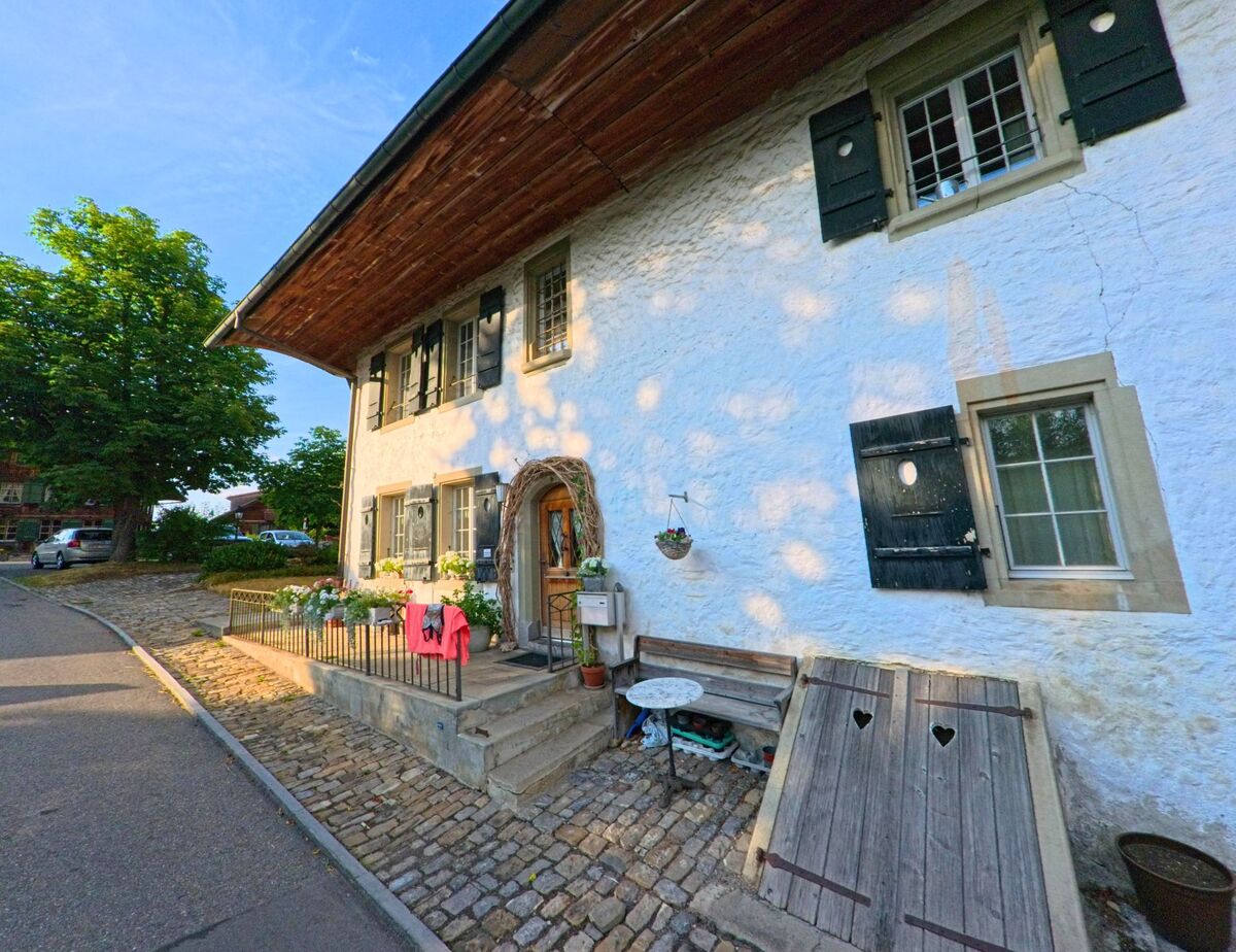 In Gurzelen (3663, Kanton Bern) steht ein liebevoll gepflegtes Haus mit blauen Fensterläden und dekoriertem Eingangsbereich an einer Kopfsteinpflasterstrasse.