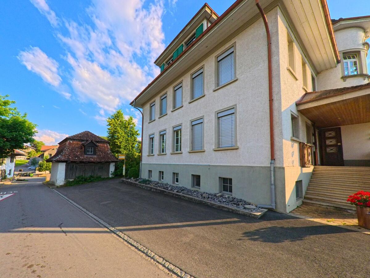 In Gurzelen (3663, Kanton Bern) steht ein stattliches Gebäude mit breiter Eingangstreppe an einer ruhigen Quartierstrasse, daneben ein kleiner Holzschopf mit Ziegeldach.