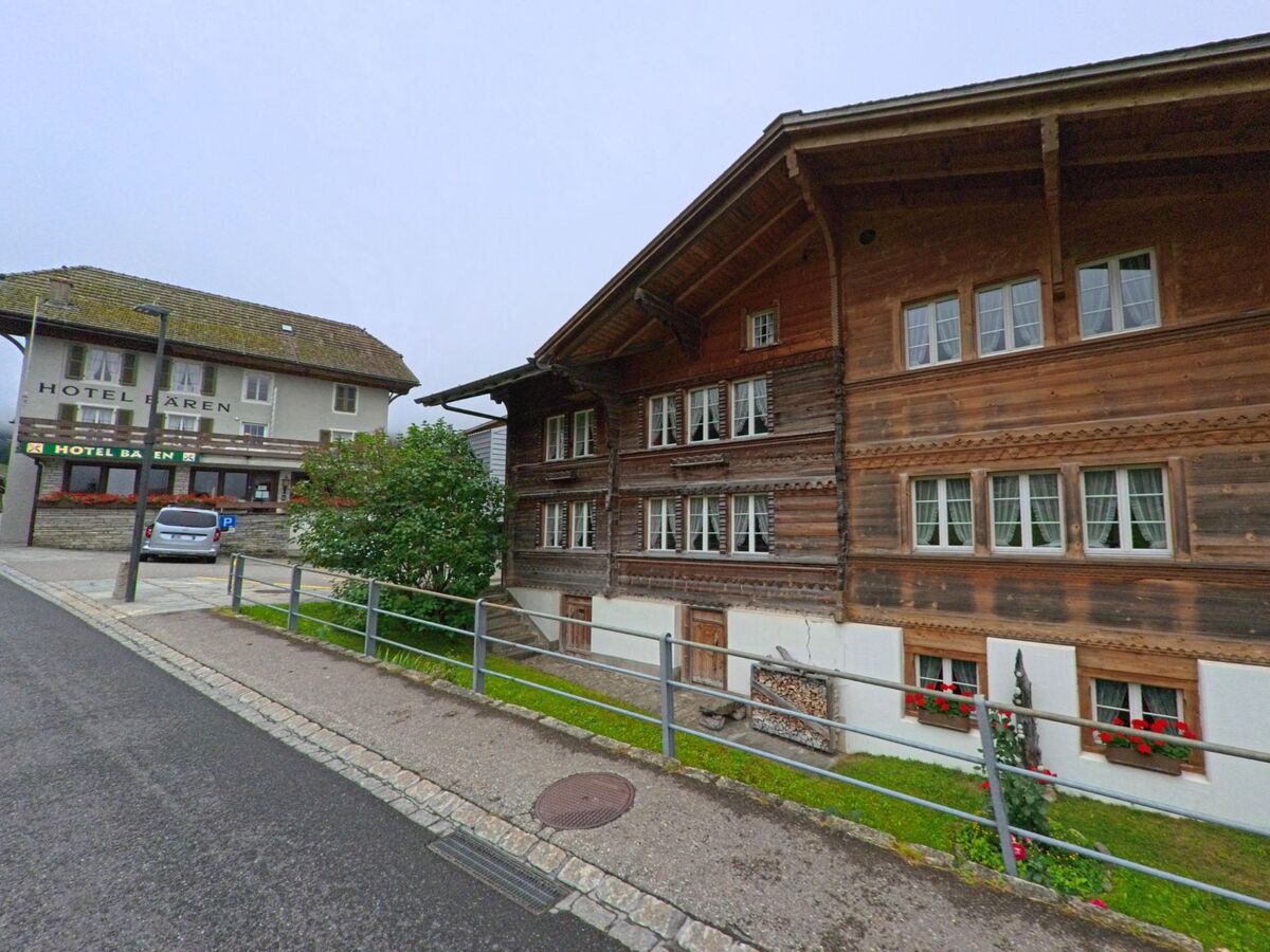 In Guttannen (3864, Kanton Bern) steht ein traditionelles Berner Oberländer Holzhaus mit roten Blumenkästen gegenüber dem Hotel Bären an einer leicht ansteigenden Strasse.