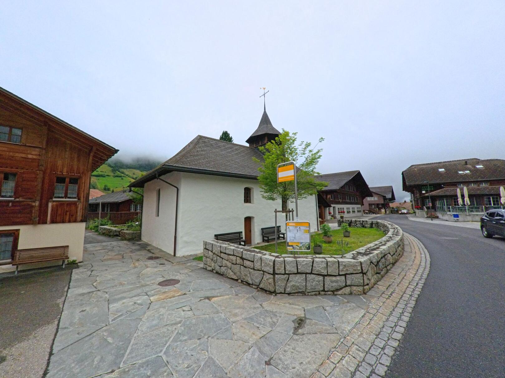 In Guttannen (3864, Kanton Bern) steht eine kleine Kirche mit markantem Turm zwischen traditionellen Holzhäusern und einer Postautohaltestelle bei bewölktem Himmel.
