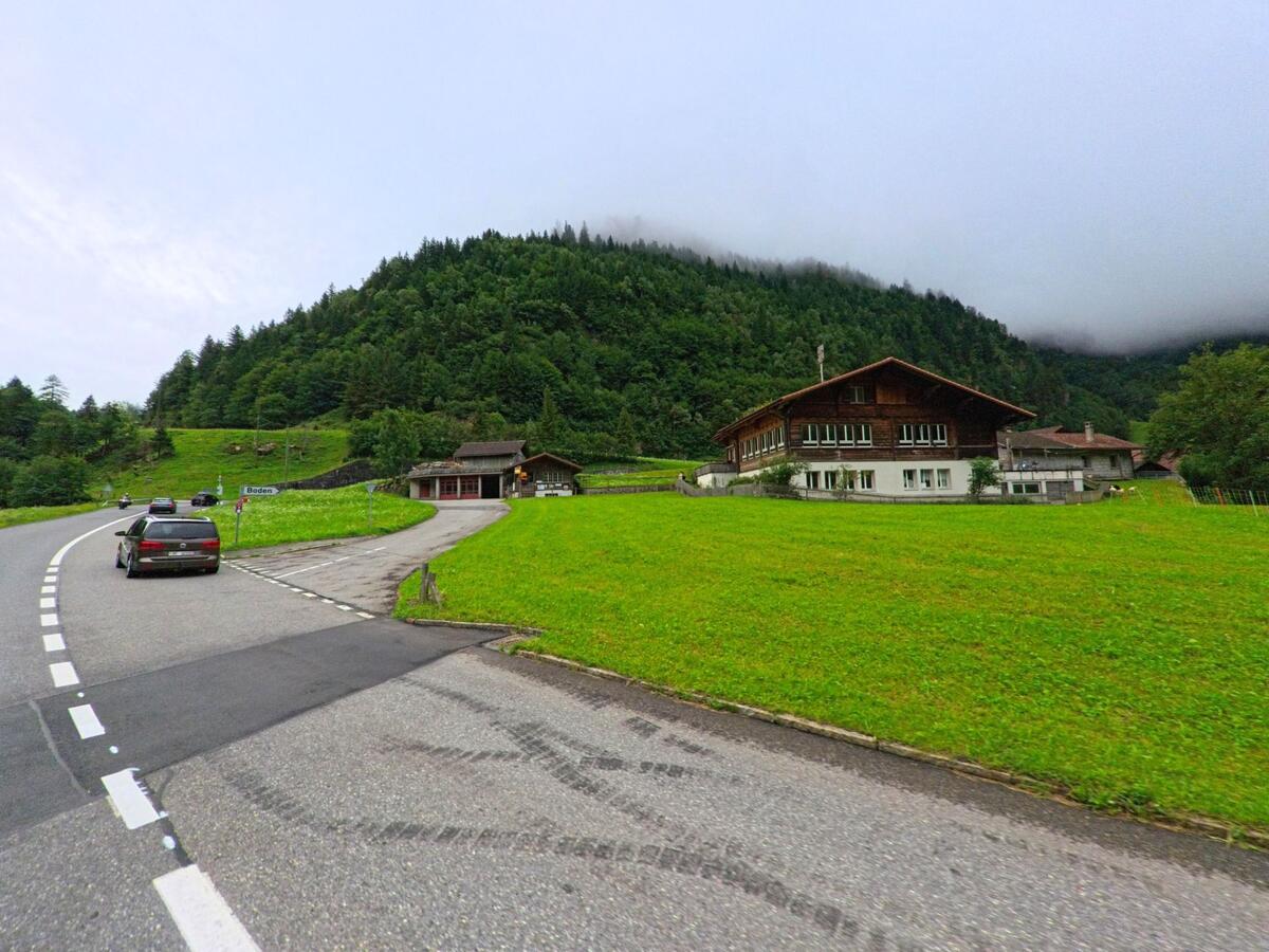 Am Ortseingang von Boden bei Guttannen (3864, Kanton Bern) liegt ein grosses Holzhaus auf einer grünen Wiese unterhalb eines bewaldeten Berghangs mit tief hängendem Nebel.