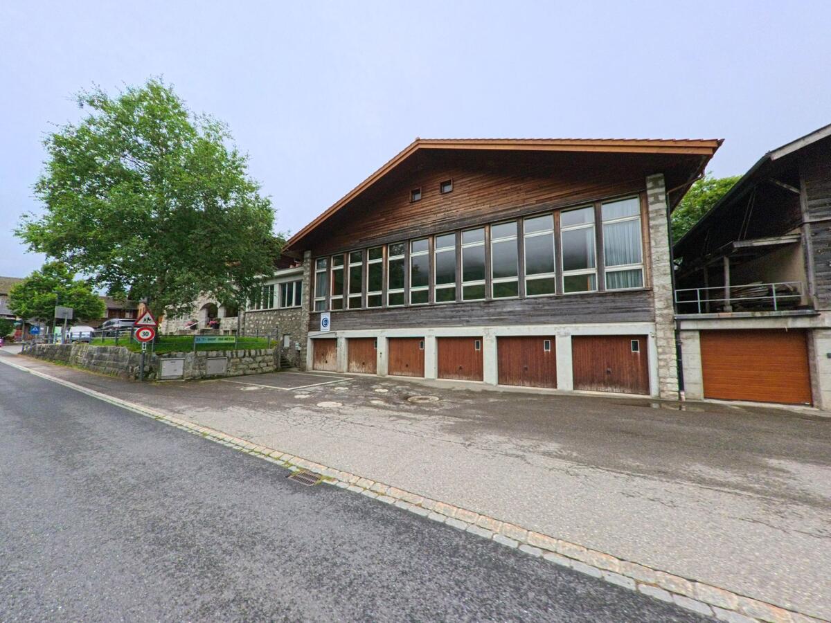 Das Schulhaus in Guttannen (3864, Kanton Bern) besitzt eine lange Fensterfront und mehrere Garagentore, eingerahmt von einem Baum und Verkehrszeichen.