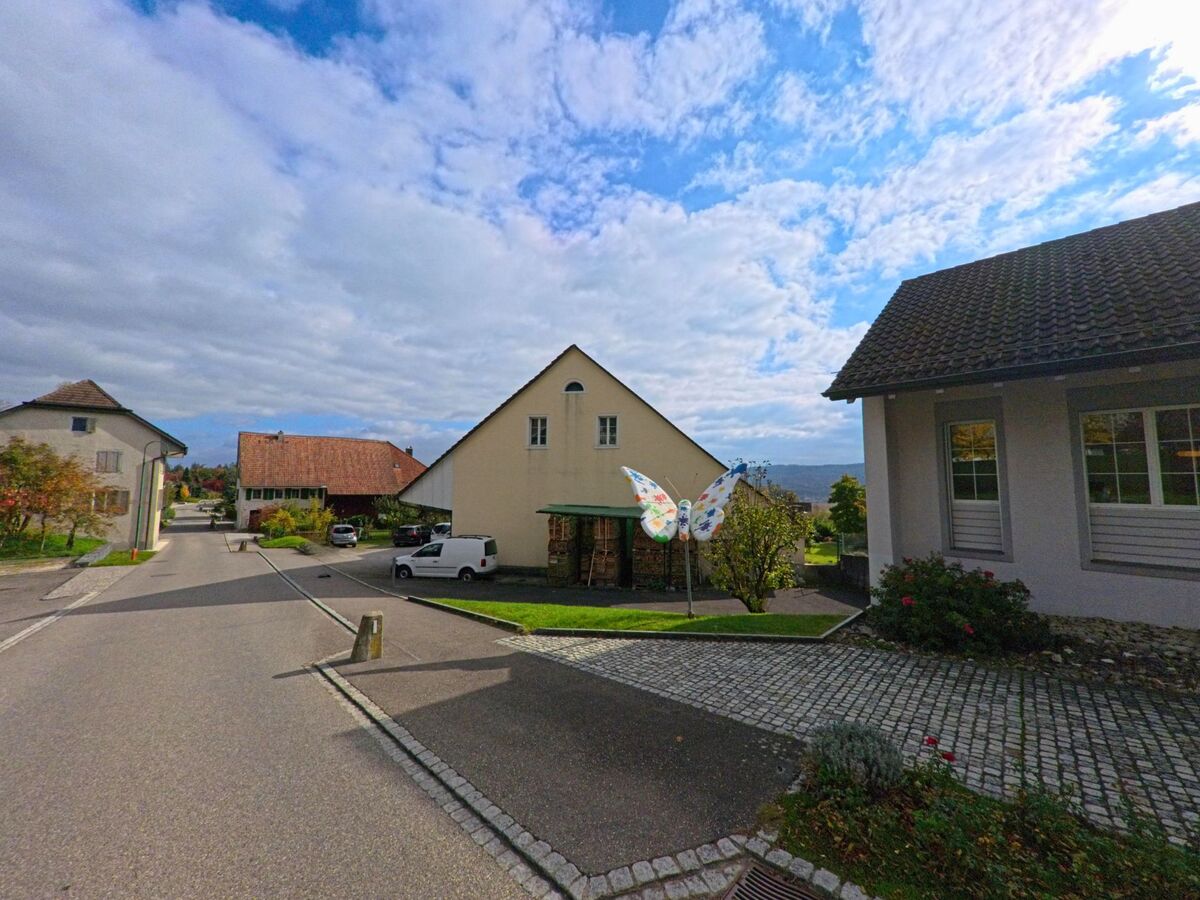 In Habsburg (5245, Kanton Aargau) steht ein helles Haus mit blauen Fensterläden neben einem öffentlichen Anschlagbrett an einer ruhig verlaufenden Strasse.