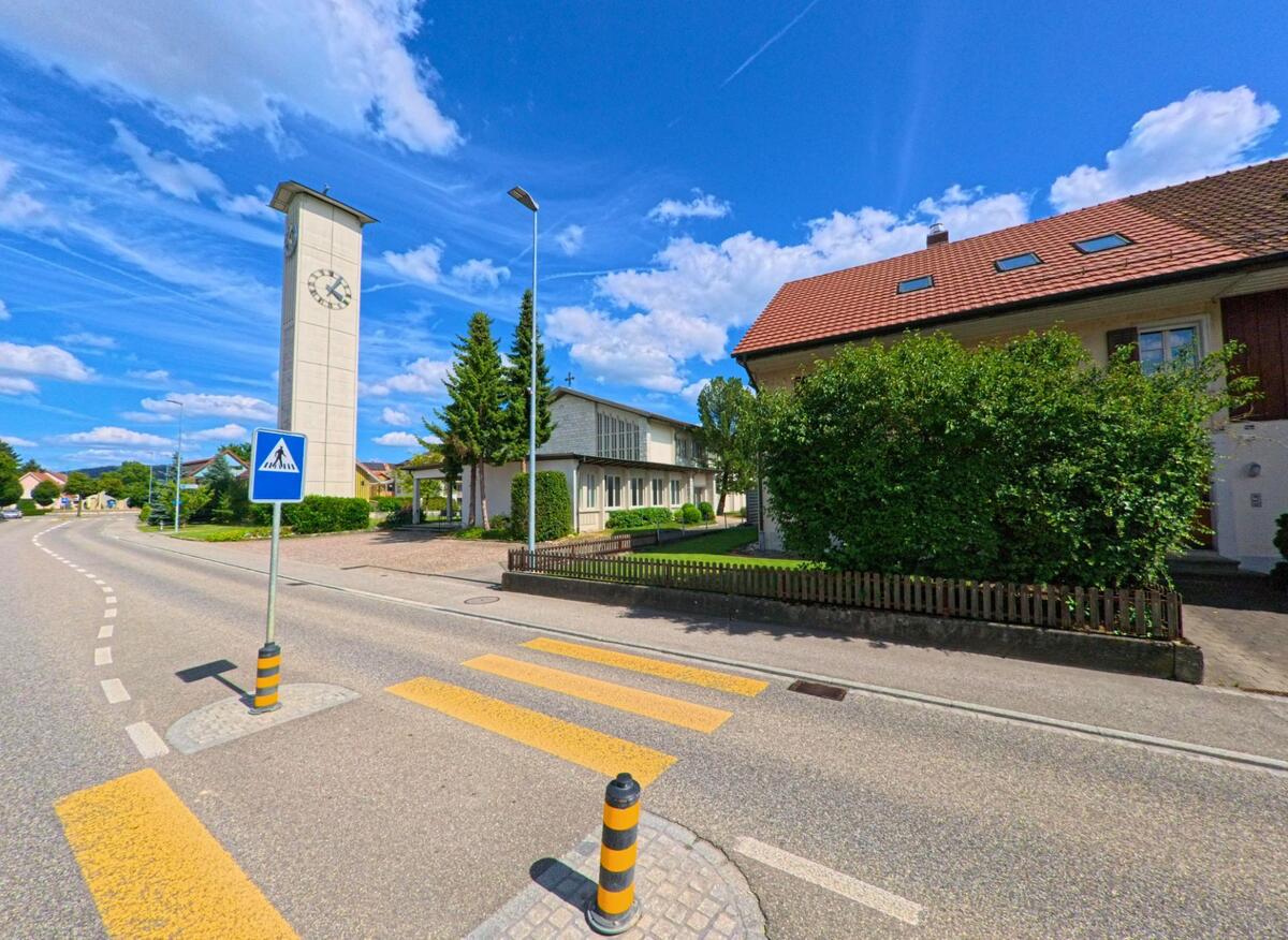 In Härkingen (4624, Kanton Solothurn) steht eine Kirche mit markantem Uhrturm direkt an einer Hauptstrasse mit Fussgängerstreifen und angrenzender Wohnbebauung.