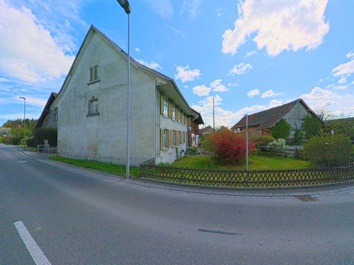 Ein älteres Wohnhaus mit gepflegtem Vorgarten steht an einer Strasse in Hagenbuch im PLZ-Gebiet 8523 im Kanton Zürich, umgeben von weiteren ländlichen Gebäuden.