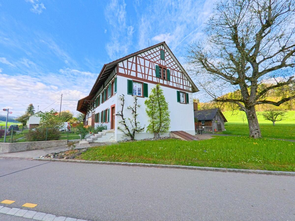 Ein sorgfältig gepflegtes Fachwerkhaus mit grünen Fensterläden steht in Hagenbuch im PLZ-Gebiet 8523 im Kanton Zürich am Rand einer Strasse, umgeben von Obstbäumen und Wiese.