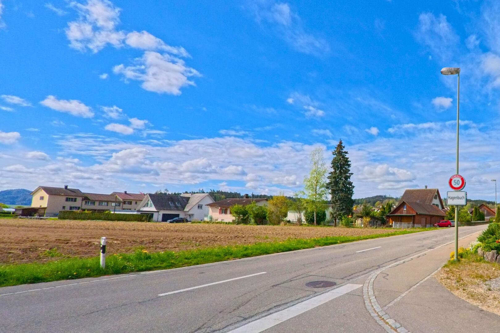 Am Ortseingang von Hagenbuch in der PLZ 8523 im Kanton Zürich verläuft eine Landstrasse entlang von Wohnhäusern und Feldern unter einem weiten Himmel mit lockerer Bewölkung.