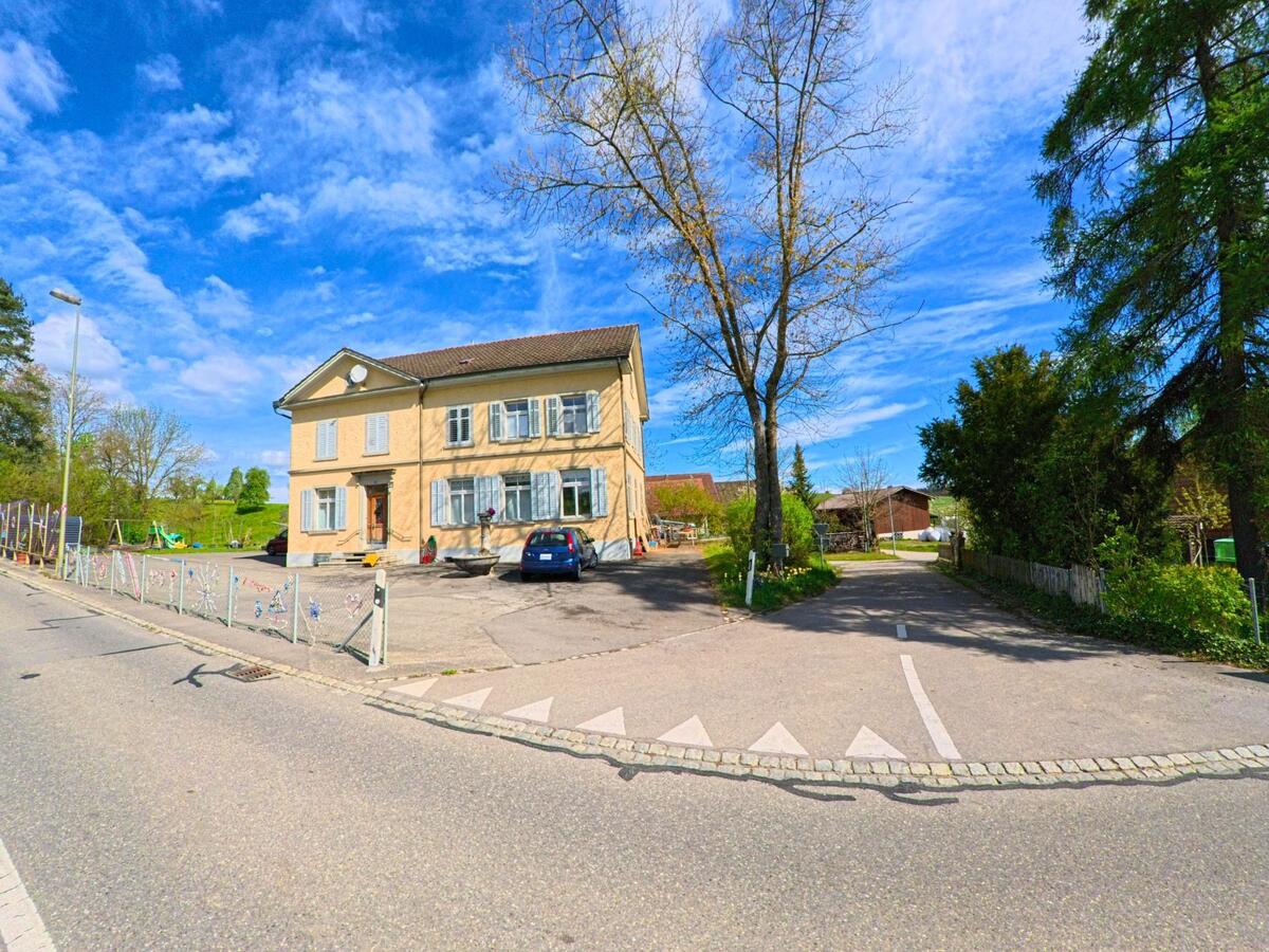 Ein helles Schulhaus mit Spielplatz in der Gemeinde Hagenbuch im PLZ-Bereich 8523 im Kanton Zürich steht an einer verkehrsberuhigten Strasse, umgeben von Bäumen und Wiesen.